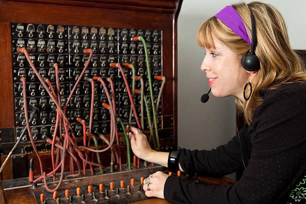 An obviously staged stock photo of a woman with a goofy grin on her face, who was not even born when the last manual switchboard was taken out of service, sitting at a manual phone switchboard. She’s wearing a purple headband and a modern-ish headset that would be completely incompatible with the switchboard. The switchboard has many of its cords plugged into the plugboard.