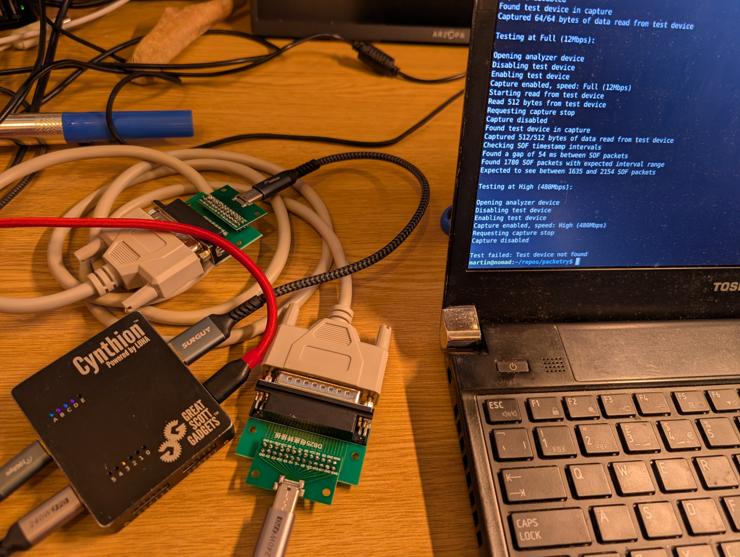 A messy desk with some DIY USB-C to DB25 parallel adapters, a Cynthion testing device and a laptop displaying a successful 12Mbps packet test