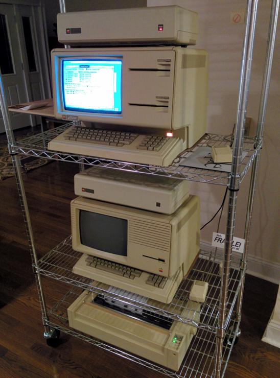 A wire rack holding an Apple Lisa 1 computer (currently operating, with its screen on) and below that, an Apple Lisa 2/10 computer. At the bottom is a wide-carriage Apple Imagewriter dot-matrix printer.